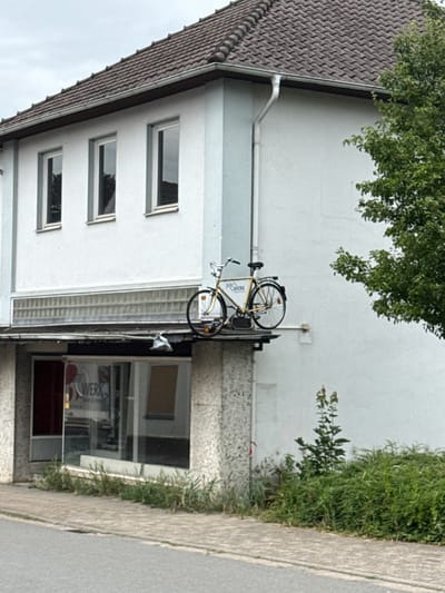 This image captures a building facade in Bassum, Germany. The building has a white upper story with multiple rectangular windows and a dark brown tiled roof. A yellow bicycle is mounted on a metal rack attached to the building's exterior wall, above what appears to be a storefront or entrance with large glass windows. The bicycle is positioned vertically. Green foliage, including tree branches and bushes, frames the scene on the left and right sides. The ground in front of the building is paved with light-colored concrete slabs. A sidewalk runs alongside the building. The overall impression is one of an urban or semi-urban setting. The lighting suggests it could be daytime, with overcast skies indicated by the soft, diffused light. There is some visible text on the building's glass facade, including the word "WERK" which is likely part of a business name. The image is oriented vertically.