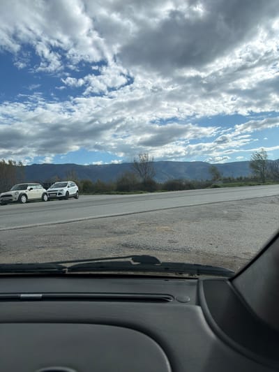 The image shows a view from inside a vehicle, looking out over a road with two parked cars. In the background are mountains and a partly cloudy sky. The location is likely near the city of Turnovo, North Macedonia given the geography and context. A portion of the vehicle's interior is visible on the right side of the frame.