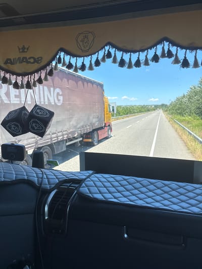 The image is taken from the inside of a truck cab, looking through the windshield. A beige sun visor with "SCANIA" logo and decorative tassels hangs across the top of the windshield. Two black dice with the Scania logo hang from the visor. In front of the windshield, two black dice-shaped air fresheners are hanging. A truck with a red trailer is driving on the highway ahead. The highway is flanked by green trees and a guardrail. The sky is blue with scattered clouds. The foreground shows the interior of the truck cab, with a quilted dashboard cover and a black dashboard. This scene appears to be on a highway in or near Κορνοφωλιά, Greece.