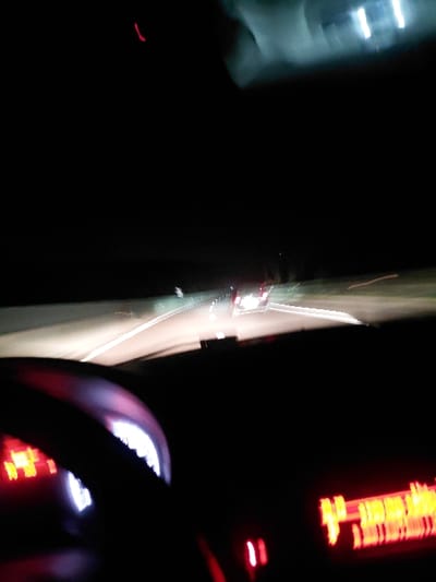 A blurry, dark image appears to be taken from inside a moving vehicle at night. The dashboard is visible with illuminated gauges and displays, reflecting red and white light. Outside, the road is a streak of light, suggesting speed and motion. Headlights from other vehicles are visible as blurred points of light. The overall impression is one of fast-paced travel through darkness.