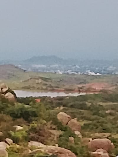 This is a blurry, aerial view of a rocky, hilly landscape with vegetation and a body of water. In the distance, a hazy cityscape is visible, suggesting an urban or suburban area within a natural setting. The image appears to be taken from a high vantage point, possibly a hill or drone, overlooking a wide expanse of land. The foreground features large rocks and green foliage, while the midground shows a calm body of water reflecting the overcast sky. The background is a blurred panorama of hills and the distant urban settlement. Without further information or clearer detail, it is impossible to ascertain the specific location within Gero, Nigeria, or to identify any specific people, subjects, objects, or actions. The environment is predominantly natural, characterized by hills, rocks, and vegetation, with the presence of a distant human-made settlement.