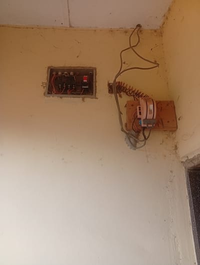 The image depicts an interior corner of a room with light yellow walls and a white ceiling, showing signs of dust and cobwebs. On the left wall, a recessed electrical panel box is visible, containing several circuit breakers with red and black wires. To the right of the panel, a separate electrical device, possibly a fuse holder or a junction box, is mounted on a wooden plank attached to the wall. This device is pink and white, with a gray lever. Several electrical wires, some coiled, are connected to both the panel and the separate device, and run along the wall and ceiling. The overall scene suggests an area with exposed electrical wiring and a need for maintenance or a cleanup. The location is identified as Zanzi, Nigeria.