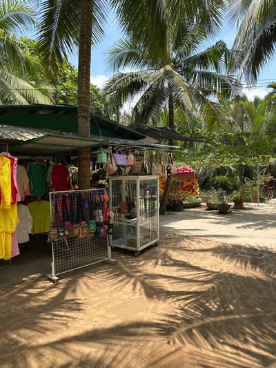 The image captures a street scene in Tân Thạch, Vietnam, featuring a small vendor's stall. The stall is displaying various merchandise, including brightly colored clothing items (yellow, pink, red, green), bags, and small souvenirs on a wire grid. A glass display case sits next to the grid. Above the stall, assorted decorative flower arrangements and potted plants are visible. The scene is heavily shaded by palm trees, casting distinct shadows on the sandy ground. The sky is partially visible through the palm fronds. The photograph is oriented sideways, with the sky appearing to be on the left.