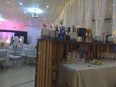 The image shows a bar area set up in a large room, likely for a wedding reception, in Lindi Street, Tanzania, located at coordinates -3.347 latitude and 37.332 longitude. A wooden bar constructed from wooden crates, with a white tablecloth, holds a variety of alcoholic beverages including bottles of wine, spirits, and a large glass pitcher of punch. The room is decorated with white curtains and white string lights. There are chairs and tables set up in the background, along with a stage set up with floral arrangements. A man dressed in a white shirt and dark pants is visible in the background.