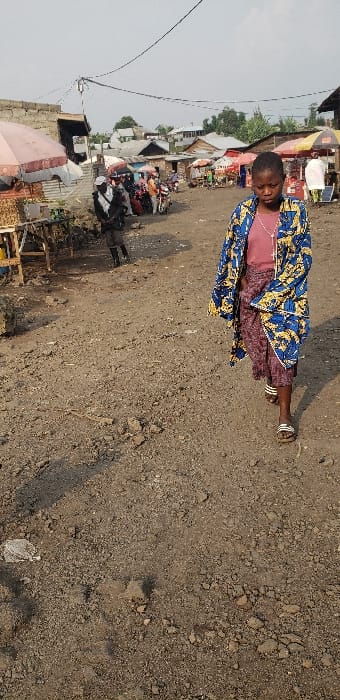 The image depicts a daytime outdoor scene in what appears to be a market or commercial area within Buzove, Democratic Republic of the Congo. The foreground is dominated by a young female, approximately early to mid-teens, walking towards the viewer on an unpaved, uneven dirt and gravel ground surface. She is wearing a long-sleeved, knee-length outer garment with a blue, yellow, and white patterned design over a maroon or reddish-brown top and a patterned maroon or purple skirt. Her feet are shod in white and black striped slide sandals. Her head is slightly bowed, and her gaze is directed downwards.

In the mid-ground and background, an active street or market environment is visible. To the left, a vendor stall features an open pink and white umbrella, with goods displayed on a wooden table. Another individual in dark clothing and a white scarf or wrap is partially visible behind this stall. Further back, numerous people are gathered, and additional market stalls with various colored umbrellas (red, yellow) and structures are present. Several motorcycles or mopeds are parked among the people. Buildings constructed from cinder blocks or brick with corrugated metal roofs line the area. Overhead, multiple electrical wires traverse the scene against a light, overcast sky. The overall lighting suggests natural daylight, with shadows cast on the ground indicating a light source from the right side.