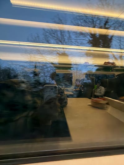 The image depicts the interior of a train carriage, viewed through a window, against an exterior backdrop of trees and sky. The location is in or near Gloucester, United Kingdom.

**Environment:** The exterior view through the window shows bare tree branches against a light blue sky, indicating either dawn, dusk, or an overcast daytime. Visible motion blur suggests the train is in transit. Inside the carriage, linear fluorescent strip lighting runs along the ceiling.

**Subjects & Objects:**
*   **Foreground (reflection):** A person's hand, presumably the photographer, is visible holding a silver-colored smartphone with a prominent dual or triple camera array, indicating a contemporary model (e.g., iPhone 11/12/13 series). This reflection obscures parts of the interior view.
*   **Interior (visible and reflected):**
    *   A light-colored, rectangular table or tray is positioned in the immediate foreground, below the window line.
    *   To the right, on an overhead luggage rack, a brown, somewhat amorphous object, possibly a shoe or small bag, rests.
    *   Reflections within the window show other passengers. One passenger is partially visible further back on the right, wearing a light grey or white puffer jacket, holding a green mug or cup. Another figure is vaguely reflected further to the left.
*   **Actions:** The primary action captured is the taking of the photograph by the person whose hand and phone are reflected. Passengers appear to be seated and engaged in travel activities. The train is in motion.
