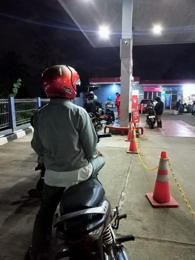 The image depicts a nighttime scene at a gas station in Cimaung, Indonesia. The primary subject is a person viewed from behind, wearing a red motorcycle helmet with "RNL" visible on it, a grey long-sleeved shirt over a white undershirt, and dark pants. They are seated on a motorcycle, appearing to be part of a queue or waiting.

In the background, under the brightly lit canopy of the gas station, several other individuals are visible, mostly on motorcycles near the fuel pumps. One person in a red uniform, possibly a gas station attendant, is standing near a pump. The station features red and white fuel pumps and a building in the background, which might be a convenience store or office, illuminated from within. A red car is partially visible near this building.

The scene is well-lit by overhead lights from the gas station canopy, contrasting with the dark night sky. Palm trees are faintly visible against the dark horizon to the left. A metal fence borders the left side of the frame. Orange traffic cones connected by yellow chains are placed on the ground, likely guiding traffic or marking a waiting area. The atmosphere appears routine, with people engaged in the common activity of refueling their vehicles at night. No distinct emotions are discernible among the individuals present. A red sign is visible on a pole near the pumps, but its text is unreadable.