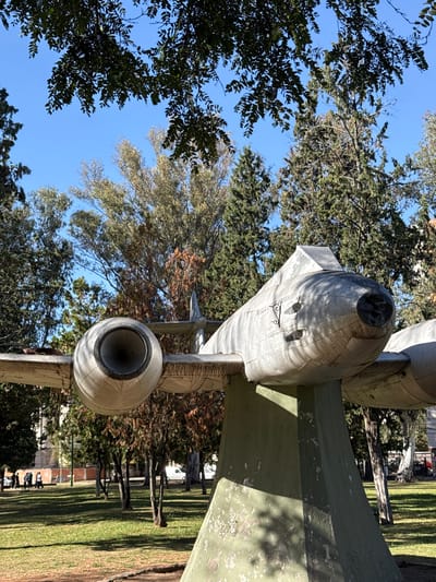 The image shows a close-up, low-angle view of a gray aircraft fuselage mounted on a support structure, likely a static display. The aircraft is surrounded by trees and lawn. The image was taken in the city of Salta, Argentina.