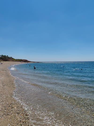 This is a photograph of a beach on a sunny day. The sky is clear blue and takes up approximately the top half of the image. The sea is a bright blue with shimmering sunlight on its surface. There are several people in the water, some swimming and some standing. The beach is made of sand and pebbles. In the background, there is a rocky coastline with some vegetation and a few palm trees. The location is likely a beach near Alexandroupolis, Greece, given the context.