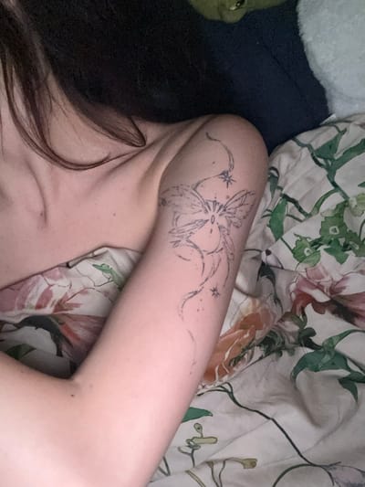 A person's arm with a floral tattoo is visible, lying on a patterned bedsheet. The tattoo is of a flower with trailing lines ending in small stars. The person's dark hair is visible near their shoulder. The bedsheet has a floral pattern with green, red, and white colors. A plush toy is partially visible on the left side of the image, near the person's head.