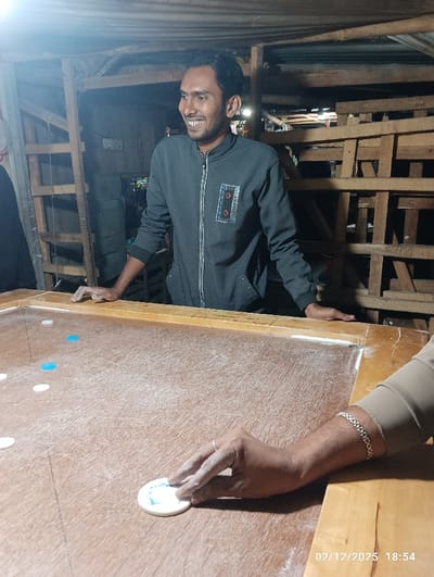 An indoor scene in Maliat, Bangladesh, depicts three individuals engaged in a carrom game on 2025-02-12 at 18:54. The foreground features a light-brown wooden carrom board with scattered white and light blue carrom men and a white striker. A right arm and hand, wearing a light brown long-sleeved garment, a silver-colored chain bracelet, and a silver-colored ring on the ring finger, are positioned in the bottom right, with the hand grasping the white striker, coated in white powder. At the opposite side of the board, a male individual with short dark hair, dark stubble, and a smiling expression stands leaning forward with both hands resting on the board's edge. He wears a dark grey zippered jacket with a rectangular, multi-colored embroidered patch on the left chest. Another person's hand is visible near his right hand on the board. The background consists of a rustic interior with exposed wooden framing and shelving, suggesting a simple structure, possibly a workshop or casual gathering area. A single overhead light source illuminates the scene, casting shadows.