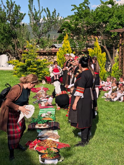 The image shows a gathering of people in what appears to be a park or garden, possibly in Sapareva Banya, Bulgaria. Several women are dressed in traditional Bulgarian clothing. A woman wearing a brown cardigan and red plaid skirt is holding a white cloth and bending over a line of food items that are placed on top of fabric laid on the grass. Another woman in traditional attire is kneeling near the food. Several other women in similar traditional costumes are standing nearby. In the background, children are seated on the grass. Trees and buildings are visible in the background. The scene appears to be part of a cultural event or ceremony.
