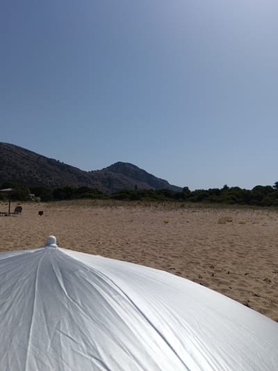 The image shows a wide, sandy beach on a clear, sunny day. In the foreground, a large, silver beach umbrella is visible, partially obscuring the view of the sand. The sand is light brown and appears undisturbed, with some scattered debris. In the middle ground, there is a line of green bushes and trees, with some low buildings and two empty lounge chairs beneath a straw umbrella visible on the left side. Beyond the vegetation, a large, rocky mountain range dominates the background, with a clear blue sky overhead. The scene appears to be a tranquil coastal landscape in the Municipal Unit of Larissos, Greece.