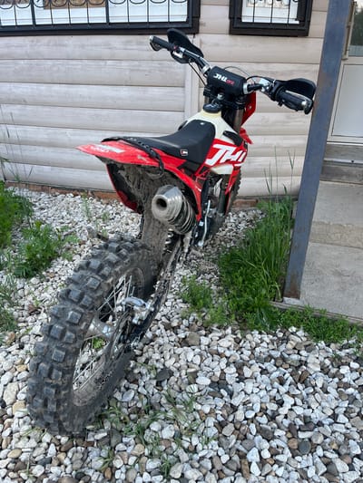The image shows a dirt bike parked on a gravel surface next to a wooden building. The bike is red and white, with knobby tires, indicating it is designed for off-road use. The rear tire is partially visible, showing its large tread pattern. The bike is dirty, suggesting it has recently been ridden. In the background, the wooden building appears to be a house or outbuilding, with windows visible. The environment is outdoors, with grass and weeds growing around the gravel. The location is identified as Krasnyi Yar, Russia. There are no people visible in the image.