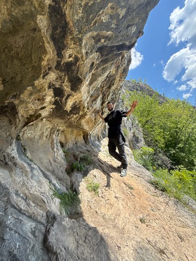 A man wearing dark clothing and light sneakers is positioned horizontally between two rock walls, appearing to either climb or fall between them. The location is characterized by a narrow crevice formed by the rock walls, with some greenery visible at the top of the frame. Above, a blue sky with scattered white clouds is present. The scene appears to be in a natural environment, not a city, so the location in Lipnitsa, Bulgaria may not be the true location.