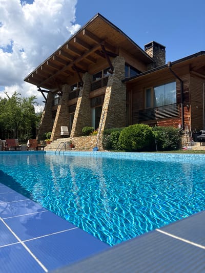 The image depicts a modern, multi-story wooden and stone house with a prominent, steeply pitched roof and exposed wooden beams. A bright blue swimming pool occupies the foreground, its surface rippling under sunlight. The house features large windows, a stone chimney, and is surrounded by lush green trees and manicured bushes. A set of outdoor chairs and loungers are visible near the pool. Cleaning tools, including a broom and a pool skimmer, are propped against the house. The scene is set in Dobrinishte, Bulgaria, under a partly cloudy blue sky. There are no people visible in the image.
