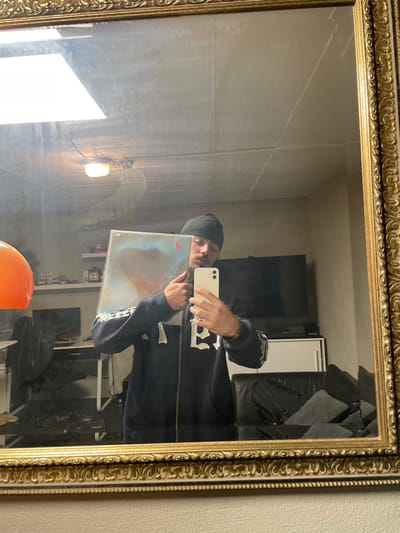The image depicts an indoor scene in Oberhausen, Germany, reflected in a large, ornate gold-framed mirror. An adult male, wearing a black beanie and a dark blue zip-up hoodie with white graphic designs on the sleeves and chest, stands centrally, holding a white smartphone in his right hand to capture the mirror selfie. His face, partially obscured by the phone and a rectangular item held in his left arm, shows visible facial hair including a mustache and goatee. The item he holds is a framed artwork or album cover featuring abstract blue, brown, and white tones, with a red sticker visible on its upper left corner.

The reflected environment reveals a room with light-colored walls and a paneled ceiling from which two light fixtures (one large square, one smaller circular) are suspended. To the left, an orange spherical object is visible next to a black desk with a monitor and a black office chair. Above the desk, a shelf displays various small items, including what appear to be model cars. To the right, a large flat-screen television is visible above a white console cabinet, next to a dark-colored sofa or armchair on which dark fabric or clothing is placed. The mirror itself exhibits some smudges or marks on its surface.