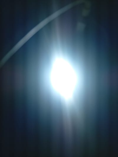 The image displays a bright light source, likely a lamp or bulb, emanating a strong glare. A faint, curved line, possibly an object reflecting light or a structural element, arcs across the top left of the frame. The background is dark and indistinct, with a subtle gradient of blues and grays, suggesting an indoor or nighttime setting. The overall impression is one of intense illumination against a shadowed backdrop. This image does not contain enough contextual information to determine its location in Mubi, Nigeria.