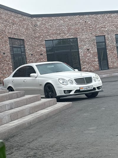 This media file shows a white Mercedes-Benz sedan parked on a wet asphalt surface next to a building with a brick facade. The car is angled, with its front facing towards the right side of the frame. A set of concrete steps is visible in the foreground, to the right of the car. The building has large, dark-tinted windows. The license plate on the car is partially visible and appears to read "OO 629 CE" with a Russian flag emblem. The overall impression is of an overcast day, given the wet surfaces and dull lighting, suggesting it might be raining or has recently rained. The setting appears to be an urban or semi-urban area in Gizel, Russia, as indicated by the license plate's potential origin and the provided location context. No people are visible in the frame, and the scene is static, with the car parked.