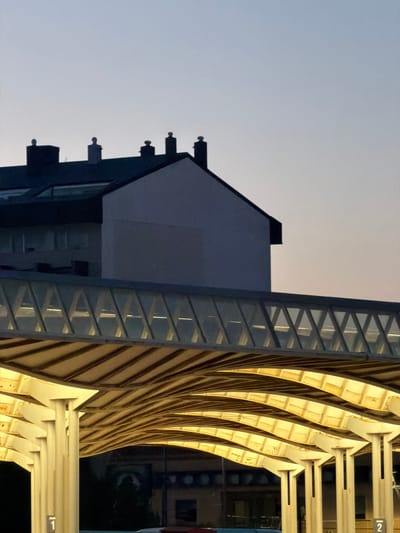 The image shows a modern architectural structure with a curved, yellow-lit roof supported by cream-colored pillars. Above this structure, a pedestrian walkway with a translucent, diamond-patterned roof is visible. Further up, a building with a dark sloped roof and white facade is silhouetted against a gradient twilight sky. Several chimney pots with rounded tops are positioned along the roofline of the building. The scene appears to be a transportation hub or a public space, possibly a train station, given the numbering signs (labeled "1" and "2") visible on some of the pillars in the foreground, suggesting platforms or entrances. The lighting suggests it is dusk or early dawn. The location is identified as Castelló De La Plana, Spain.