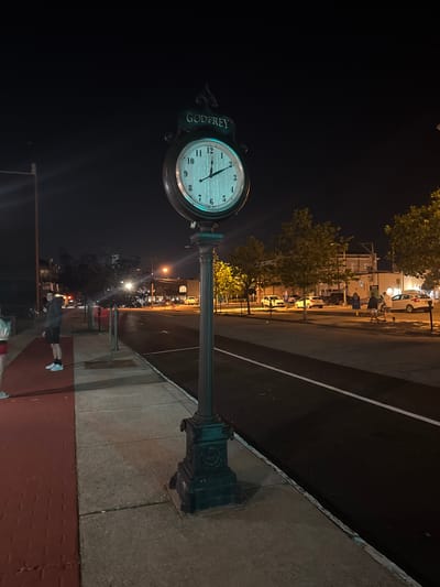 An eye-level, full-shot photograph captures a street scene at night. The prominent subject is a dark green, ornate street clock with a white face and black numerals, topped with the word "GODFREY" in raised lettering. The clock's hands indicate the time is approximately 1:45. The clock stands on a dark, decorative metal post with a square base. To the left of the clock, a sidewalk is visible, transitioning from a red brick-like surface to a gray concrete one. A person in athletic wear, a grey jacket and black shorts, stands facing the camera on the gray concrete section of the sidewalk. Further down the sidewalk, several trees with sparse foliage are illuminated by streetlights. Across the street, a road with white lane markings is lined with parked cars and buildings. The overall environment suggests a public area in a town or city at night. The location appears to be Ocean City, United States of America, specifically on a street with businesses and pedestrian areas.