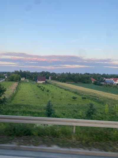 The image depicts a rural landscape with scattered houses and cultivated fields under a pale blue sky with wispy pink clouds. In the foreground, a light-colored metal guardrail runs horizontally across the bottom of the frame, suggesting the viewpoint is from a road or elevated position. Beyond the guardrail, a lush green field stretches into the middle ground, dotted with small trees and what appear to be garden plots. A single house with a dark roof is situated in the middle of this field. Further in the distance, more houses with red and grey roofs are visible, nestled amongst trees. A field of yellowing crops, likely wheat or barley, is also present. The overall scene conveys a peaceful, suburban or rural setting in Podłęże, Poland. No people are visible in the image.