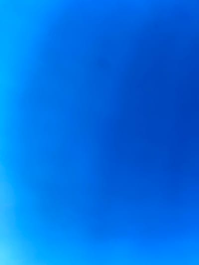 This is a full-frame, high-angle, outdoor shot of a clear blue sky. There is a subtle gradient of color, with the sky appearing lighter on the left side of the frame and a deeper blue towards the right and top. No objects or people are visible in the frame. The image depicts the sky over the village of Kaliuzhyntsi, Ukraine.