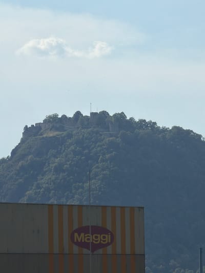 The image shows a view of Hohentwiel castle ruins on a forested hill in Singen (Hohentwiel), Germany. In the foreground, a building with a large sign displaying the "Maggi" logo is visible. The sky is a pale blue with a few wispy clouds. The scene appears to be a sunny day.