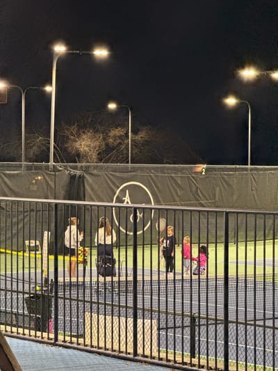A nighttime shot reveals an outdoor tennis court viewed through a fence. Players are visible on the illuminated court surface. A backdrop behind the court features a large circular logo. To the left, a line of streetlights illuminates bare tree branches against a dark sky. The scene suggests a nighttime tennis practice or match.