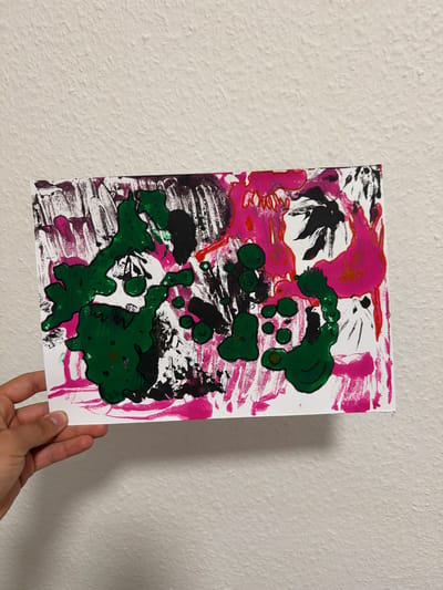 A hand holds a rectangular piece of paper with an abstract painting on it, which is leaning against a textured white wall. The painting features splatters and broad strokes of various colors, including vibrant pink, deep green, black, and white. Some of the paint is applied thickly, creating a textured effect. The background wall is a neutral white with a rough, stucco-like finish, and a soft shadow is cast on the wall behind the paper. The lighting appears to be artificial and diffused, illuminating the artwork without harsh shadows. The image does not contain any elements that would indicate a specific location in Nienburg/Weser, Germany.