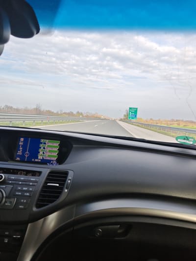 The image depicts the interior of a vehicle in motion on the A1 Maritsa Motorway, near Trilistnik, Bulgaria, looking forward through the windshield under an overcast sky. The dashboard is dark gray, featuring a central infotainment screen displaying a navigation map with a route, current time (16:50), 246 km remaining, and an estimated arrival time of 18:46. The screen also notes "A1, Maritsa." Below it are car audio and climate controls.
The highway is multi-lane, with a metal guardrail separating the direction of travel from the opposite lanes. On the right side of the road, a large green highway sign indicates exits for "Svilengrad," "Plovdiv," "Burgas," and "Sofia," specifying an upcoming exit for "Radinovo / Rakovski" in 500 meters. The surrounding landscape consists of flat fields and sparse, bare deciduous trees, suggesting a late autumn or early spring season. No individuals are visible. The windshield shows minor dust or debris specks, and a green sticker is affixed to the lower right portion of the glass.