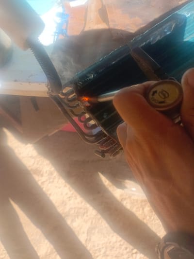 In La Cruz del Pastel, Venezuela, a close-up image shows a person's hand holding a welding torch to repair a copper coil of an air conditioning unit. The torch flame is visibly hot, and a small amount of smoke is rising from the contact point. The background is blurred but suggests an outdoor or workshop environment with debris and sunlight casting shadows. The person's right hand is the primary focus, with the left side of the frame showing a portion of the air conditioning unit's fins and tubing.