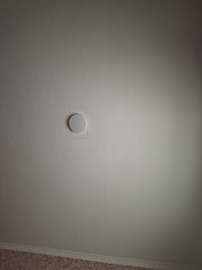 The image shows a white, circular device mounted on a textured, light-colored ceiling. The device is centrally located in the frame, with a slight shadow cast on its right side, suggesting a light source from the left. The ceiling itself appears to be painted or wallpapered, exhibiting a uniform, slightly bumpy texture. A decorative cornice is visible along the bottom edge of the frame, separating the ceiling from what appears to be a patterned carpet or floor covering below. No people or animals are present in the image. The scene is devoid of any context that would specifically place it in Khujand, Tajikistan.