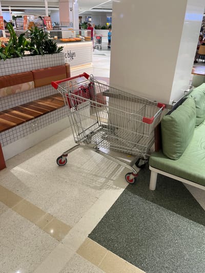 The image captures an indoor scene within a modern shopping center, consistent with the context of Indooroopilly, Australia. The photograph is rotated approximately 90 degrees counter-clockwise.

In the foreground, a prominent silver metal shopping cart with red plastic handles and red/black wheels rests on a light-colored speckled floor. Adjacent to the cart, a green upholstered sofa with white legs and a wooden base is partially visible. The shopping cart casts distinct shadows on the floor, suggesting an overhead light source.

A large, plain white column or wall separates the immediate foreground from the deeper background. Beyond this column, the scene reveals a food court or seating area with various furnishings. These include benches with striped cushions in earthy tones (browns, oranges, and off-white) and wooden slatted benches, arranged around planters with lush green foliage. The flooring in this area continues with the light speckled tiles, interspersed with darker grey/green speckled sections and light brown decorative strips.

Several blurry figures of people are visible in the background, appearing to walk or stand, engaged in general mall activities. One person is somewhat discernible wearing a red shirt. The overall bright, even illumination suggests an indoor daytime setting, likely reliant on artificial lighting.

Visible text includes the word "Coffee" in a stylized font on a white counter or wall in the background, indicating the presence of a café or food vendor. Other text on distant displays is too small and indistinct to read. The scene depicts a typical, functional environment of a contemporary shopping mall, without any specific event or heightened activity taking place.