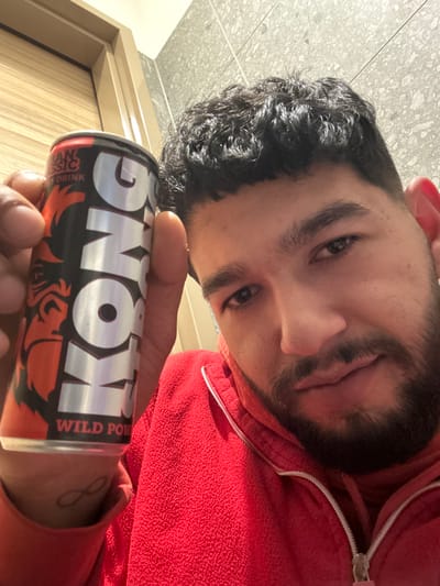 A young adult male, of apparent Mediterranean or Middle Eastern descent, is depicted in a close-up selfie taken indoors in Radovljica, Slovenia. He has dark, curly hair with a fringe, dark eyes, and a full beard and mustache. The subject is wearing a red fleece jacket with a white zipper. In his left hand, he holds a red, black, and silver "KONG Wild Power" energy drink can, prominently displaying its label which features a gorilla silhouette and the text "EUROPEAN CLASSIC ENERGY DRINK." An infinity symbol tattoo is visible on the inner wrist of his left arm. The individual is looking directly at the camera with a neutral expression. The background features light grey or beige tiled walls with visible grout lines and a partial view of a light brown wooden doorframe on the left. The scene is illuminated by indoor lighting.