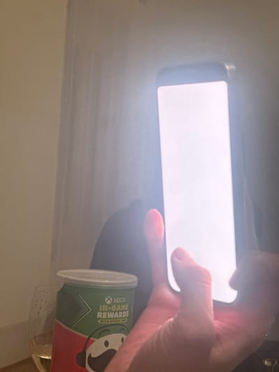 This image depicts an indoor scene, located in Roanne, France. In the immediate foreground, a human right hand holds a dark-colored smartphone vertically. The phone's screen is fully illuminated with a bright white display, serving as the primary light source. In the mid-ground, to the left of the phone, a Pringles potato chip can is visible. It features a green and red label with the Pringles character "Mr. P" wearing a red hat, and promotional text for "XBOX IN-GAME REWARDS WITH EVERY CAN." Adjacent to the Pringles can, a clear stemmed wine glass is partially visible, containing a small quantity of pale yellow liquid and exhibiting condensation droplets on its exterior surface. The background consists of a light-colored interior wall or curtain, displaying subtle vertical textures. A dark, amorphous shadow is cast behind the Pringles can and the hand, indicating an obscured figure or object. The overall environment suggests low ambient light conditions, with the phone screen providing significant illumination and contrast.