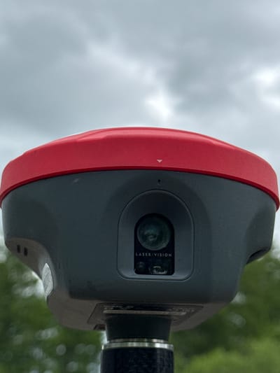 The image shows a close-up of a red and gray surveying device, possibly a total station or laser scanner, mounted on a pole. The device has a prominent red top and a dark gray body with a central lens assembly labeled "LASER/VISION." In the background, the sky is overcast with gray clouds, and blurred green foliage suggests an outdoor, possibly urban or suburban, environment. The device is positioned outdoors, and the lighting conditions indicate it is daytime. There are no people visible in the image. The location of Муртыгит, Russia is not depicted or suggested by any visual cues in the provided image.