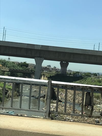 The image depicts a multi-layered urban landscape in Sandton, South Africa, under a clear blue sky.
In the immediate foreground, a grey, weathered metal barrier, likely a bridge railing, extends across the frame, with a segment of concrete roadway visible at the very bottom.
Below and beyond this railing, a heavily polluted waterway, appearing to be a river or stream, flows through a cluttered environment. The water surface and banks are densely accumulated with various forms of solid waste, including plastic bags, bottles, fabric remnants, and general refuse. On the left bank, some green vegetation is present amidst the debris.
Further into the midground, a lower, older concrete bridge or overpass with a partially visible railing crosses the waterway, spanning from the left towards the right. Behind this bridge, on the right side of the frame, a light-colored vehicle, possibly a white pickup truck or SUV, is discernible on a road.
Dominating the upper midground and background is a large, elevated concrete viaduct, likely a railway line such as the Gautrain system, supported by two prominent, robust concrete piers. Overhead power lines run parallel to and above this viaduct.
In the distant background, beyond the elevated structures, residential buildings, appearing as densely clustered houses, are visible on elevated terrain under a hazy horizon. No individuals are present in the captured scene, and no specific actions or interactions are occurring.