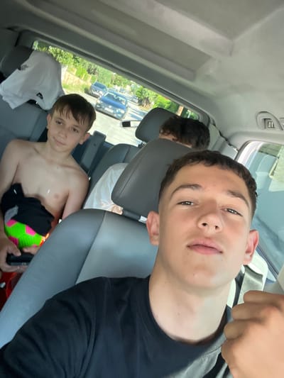 The image captures three young males inside a vehicle, appearing to be in transit. The foreground is dominated by the face of one individual, who is looking directly at the camera, with a black t-shirt visible on his torso and his right fist clenched near his chest. To his left, a second individual is seated, shirtless, holding a brightly colored ball. Further back, a third individual is partially visible, with their back to the camera. The interior of the vehicle, with its grey upholstery and ceiling, suggests it is a passenger van or similar vehicle. Outside the car windows, trees and buildings lining a street are visible, indicative of a suburban or residential area. The presence of a street with cars suggests an urban or semi-urban setting. Considering the context provided, the scene is likely located in the city of Nea Iraklitsa, Greece.