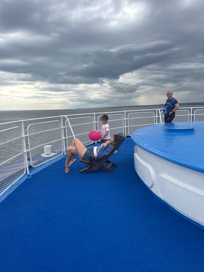 The image shows a view from the deck of a boat with a bright blue carpet. In the foreground, a woman is reclining in a lounge chair, holding a pink balloon. A young child stands nearby, looking at the woman. Further in the background, a man in a blue and black athletic shirt stands by the railing, looking out at the water. The sky is cloudy and grey, with a hint of rain in the distance. The boat appears to be on a body of water, possibly a river or a lake, with land visible in the far distance. The scene is set on a cloudy day, with the boat sailing on calm waters. The location is Ikskoe Ust'e, Russia.
