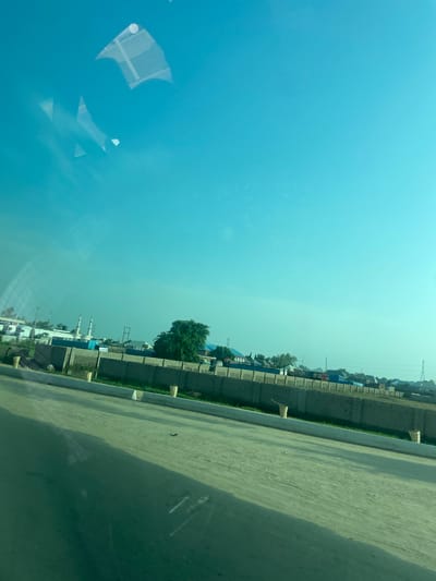 The image shows a view of Yola, Nigeria, from what appears to be a moving vehicle. The sky is a clear, bright blue, with some reflections visible on the glass. In the mid-ground, there is a long, low-rise building with a wall in front of it. Beyond the wall, there are more buildings, including what appear to be two minarets of a mosque. A large green tree is also visible. The foreground consists of a dusty, unpaved road. The overall impression is of an urban or semi-urban environment in a dry climate.