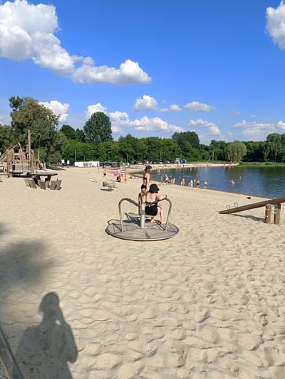 The image depicts a sandy beach area next to a body of water, likely a lake or river. The sky is blue with scattered white clouds. In the foreground, a carousel-like children's playground ride is visible, with two young individuals seated on it. To the left of the carousel, a picnic table with benches is situated on the sand. Further back, a wooden playground structure with slides and climbing elements is present. A group of people are seen swimming and relaxing in the water. The background features a dense line of trees bordering the water. In the distance, cars are parked, suggesting a public access area. The overall scene suggests a recreational day at a public beach. This location is identified as the beach area of Gubkin, Russia.