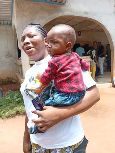 A woman stands outdoors in Umundugba, Nigeria, holding a baby. The woman has braided hair, wears a white t-shirt with a graphic, and printed shorts. She is holding sunglasses in her left hand, which has red nail polish. The baby wears a red and black patterned shirt, denim bottoms, and striped socks. The background features a building with an arched entrance where several people are standing. The ground is reddish brown. Greenery is visible to the side.