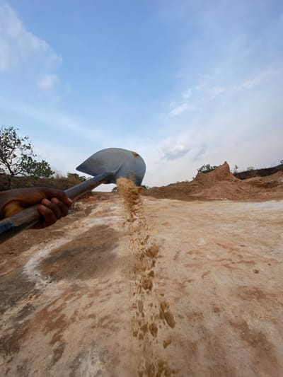 An outdoor scene in Asaga Amangwu, Nigeria, depicts an arid, excavated environment under a partially cloudy blue sky. A dark-skinned human hand, positioned in the lower-left quadrant, firmly grips the dark handle of a grey metal shovel. The shovel's blade, located centrally, is actively discharging a continuous stream of light brown sand or soil downwards and to the right, indicating material being moved. The foreground and midground consist of exposed, uneven terrain characterized by reddish-brown, tan, and white patches of earth, suggesting a construction or excavation site. In the background, rolling hills with sparse green vegetation and distinct mounds of reddish-brown earth are visible under the distant horizon.
