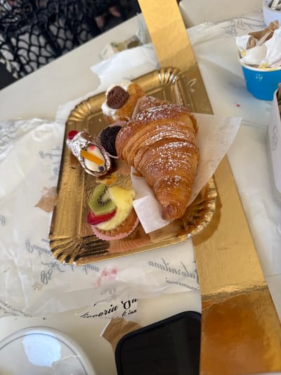 A close-up, high-angle shot shows a selection of pastries on a gold-colored tray. The pastries include a croissant dusted with powdered sugar, a cannoli topped with a red cherry and fruit, and a small tart decorated with kiwi, strawberry, and what appears to be pineapple. There are also two smaller, chocolate-covered pastries. The tray rests on a white surface covered with decorative paper that has script writing and floral motifs. In the background, a blue cup filled with crumpled paper and possibly a straw is visible. The scene suggests a treat from a bakery or café, potentially in Ardea, Italy, given the script on the paper which reads "Pasticceria da Ninetta" which is a known pastry shop in Ardea. A dark smartphone is partially visible in the lower right corner.