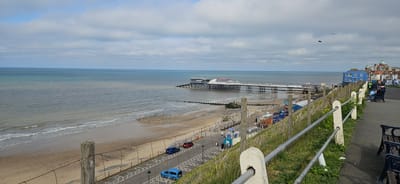 The image depicts a view of the Cromer Pier in North Norfolk, United Kingdom, from an elevated position along a paved walkway. The pier stretches into the sea, with buildings at the far end. The beach is visible below, with a mix of sand and pebbles. Along the walkway, there is a fence with wooden posts and wire, as well as metal railings. Several parked cars line the road adjacent to the beach. In the background, buildings are visible along the coastline. People are seen sitting on benches along the walkway. The sky is overcast with scattered clouds.