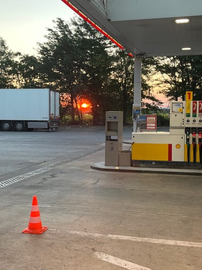 The image depicts a gas station in Gorni Dabnik, Bulgaria, at sunset. The sky exhibits hues of orange and yellow, indicating the sun is low on the horizon, partially obscured by trees. In the foreground, a bright orange traffic cone with white reflective stripes is positioned on the concrete ground. To the right, a Shell gas pump station is visible, with several nozzles attached. A white semi-trailer truck is parked to the left of the frame. The ground surface is concrete, with visible tire marks and a drainage grate running along the left side. The overall lighting suggests it is dusk.