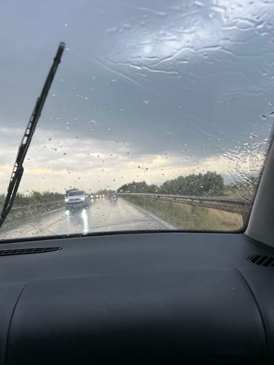The image, captured from inside a vehicle, depicts a road scene during rainfall. The windshield is streaked with water droplets and shows evidence of a windshield wiper. The road ahead is wet, with several vehicles visible, their headlights illuminated. The sky is overcast and appears to be dark with rain clouds. Lush green vegetation lines the sides of the road, suggesting a rural or semi-rural environment. The interior of the vehicle, specifically the dashboard, is partially visible in the foreground. The image does not provide any explicit indication of the specific location within Войводино, Bulgaria, only that the scene is on a road under rainy conditions.