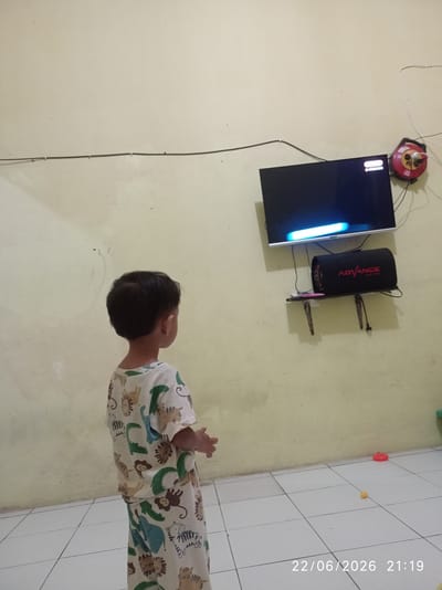 This media file shows a young child standing in front of a television screen in what appears to be a living room or bedroom. The child, facing away from the camera, is wearing pajamas adorned with a pattern of various jungle animals. The television is mounted on a wall, and below it, a black cylindrical speaker with the word "ADVANCE" printed in red is placed on a small shelf. The room has light-colored walls and a tiled floor. A red object and a small yellow ball are visible on the floor in the lower right portion of the image, suggesting a play area. The image is timestamped "22/06/2026 21:19", indicating it was taken in the evening. The overall impression is a domestic scene, likely in Sukaraja, Indonesia, where a child is engrossed in watching television. There are no clear indications of weather or strong emotions, but the scene is calm and ordinary.