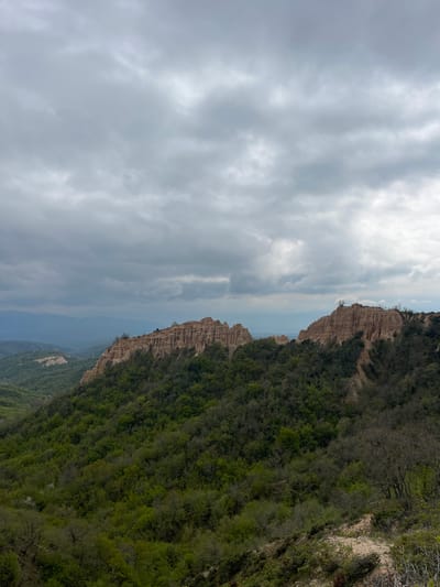 The image captures a landscape near Rozhen, Bulgaria. A rock formation with sharp peaks borders a green, tree-covered hillside. Above this formation, the sky is filled with grey clouds. The overall scene suggests a natural, elevated viewpoint, emphasizing the unique terrain and cloudy weather.